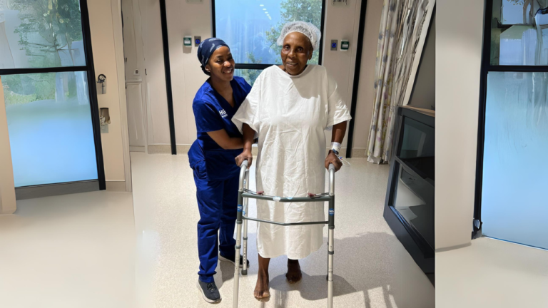 An older patient walking with a frame, supported by a nurse, soon after surgery.